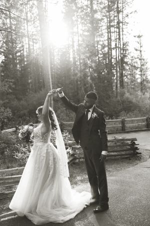 A couple shares a romantic dance moment in a scenic forest setting, with sunlight streaming through pine trees behind them.