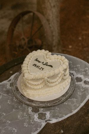A small heart-shaped white wedding cake with delicate piped frosting details sits on a silver platter with lace tablecloth beneath.