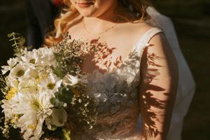 A white and natural wedding bouquet with dahlias and greenery captured in warm sunlight against a lace wedding dress.