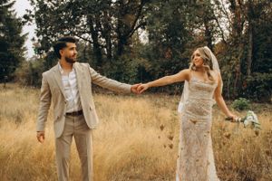 A couple in formal attire holds hands in a golden field with trees in the background during sunset.
