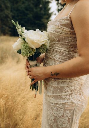 Bride in lace white dress holds elegant white rose bouquet while standing in golden wheat field.