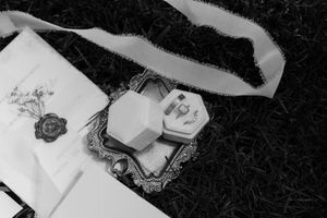 Black and white photo of wedding rings and stationery with a wax seal on dark textured background.