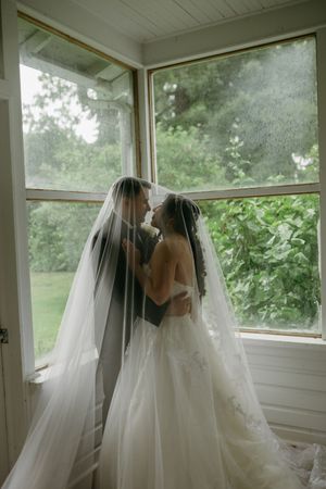 A romantic wedding moment captured by a window with flowing white veils creating an ethereal atmosphere.