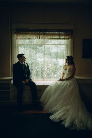 A moody wedding portrait by a window with natural light illuminating a couple in formal attire.