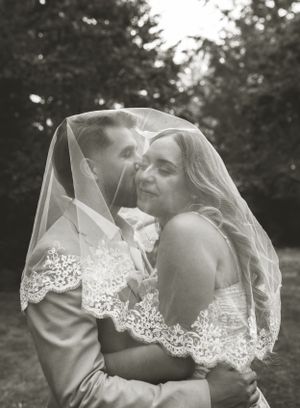 A romantic black and white wedding photo showing a tender moment under a lace veil in an outdoor garden setting.