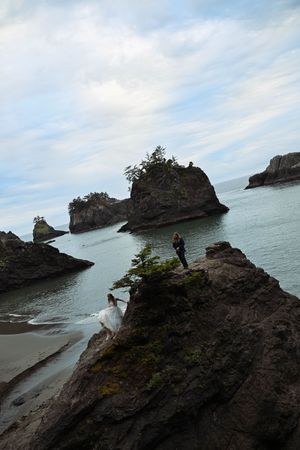 Couple in wedding attire on rocky coastal cliffs with sea stacks and calm ocean waters under a cloudy sky.