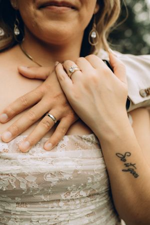 Close-up of hands wearing wedding rings against a delicate lace dress, with pearl earrings and a small wrist tattoo visible.