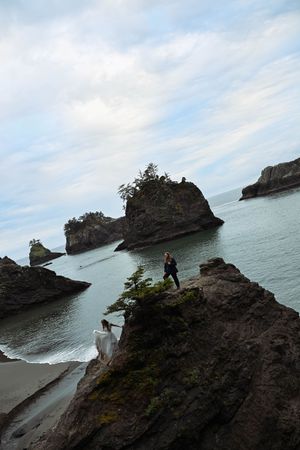Couple in wedding attire standing on dramatic rocky coastal cliffs with sea stacks and ocean views under cloudy skies.