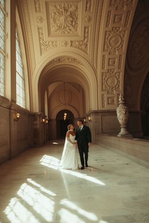A couple stands in an ornate palace hallway with dramatic sunlight streaming through tall windows onto marble floors.