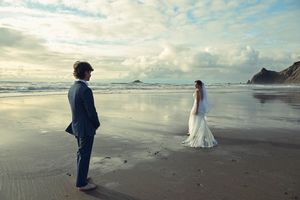A bride in a white gown and groom in a blue suit stand apart on a reflective beach with dramatic cloudy skies.