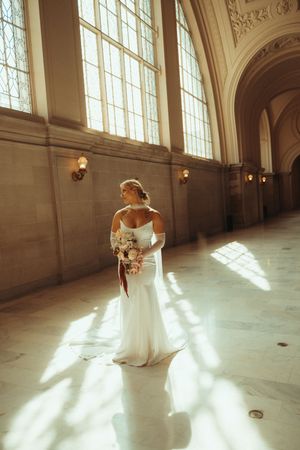 A bride in a white wedding gown twirls gracefully in an elegant sunlit hall with tall windows and ornate architecture.