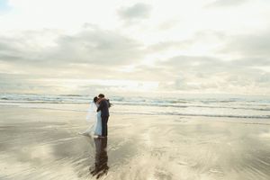 Couple embracing on a reflective beach at sunset with ocean waves and dramatic cloudy sky.