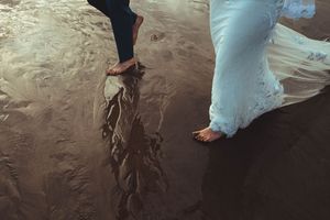 Barefoot couple walking on wet sandy beach, bride in flowing white gown with waves reflecting in the sand.