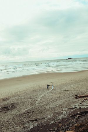 Couple in wedding attire walking hand in hand on a vast sandy beach with ocean waves under a cloudy sky.