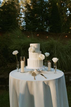 Three-tiered white wedding cake with roses displayed on round table with candles outdoors at dusk.