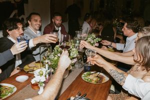 People raise their glasses in a toast during a dinner party at a rustic wooden table with plates of food and greenery decor.