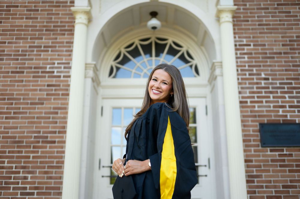 Smiling female graduate in black and gold academic regalia posing in front of a brick building with white door.