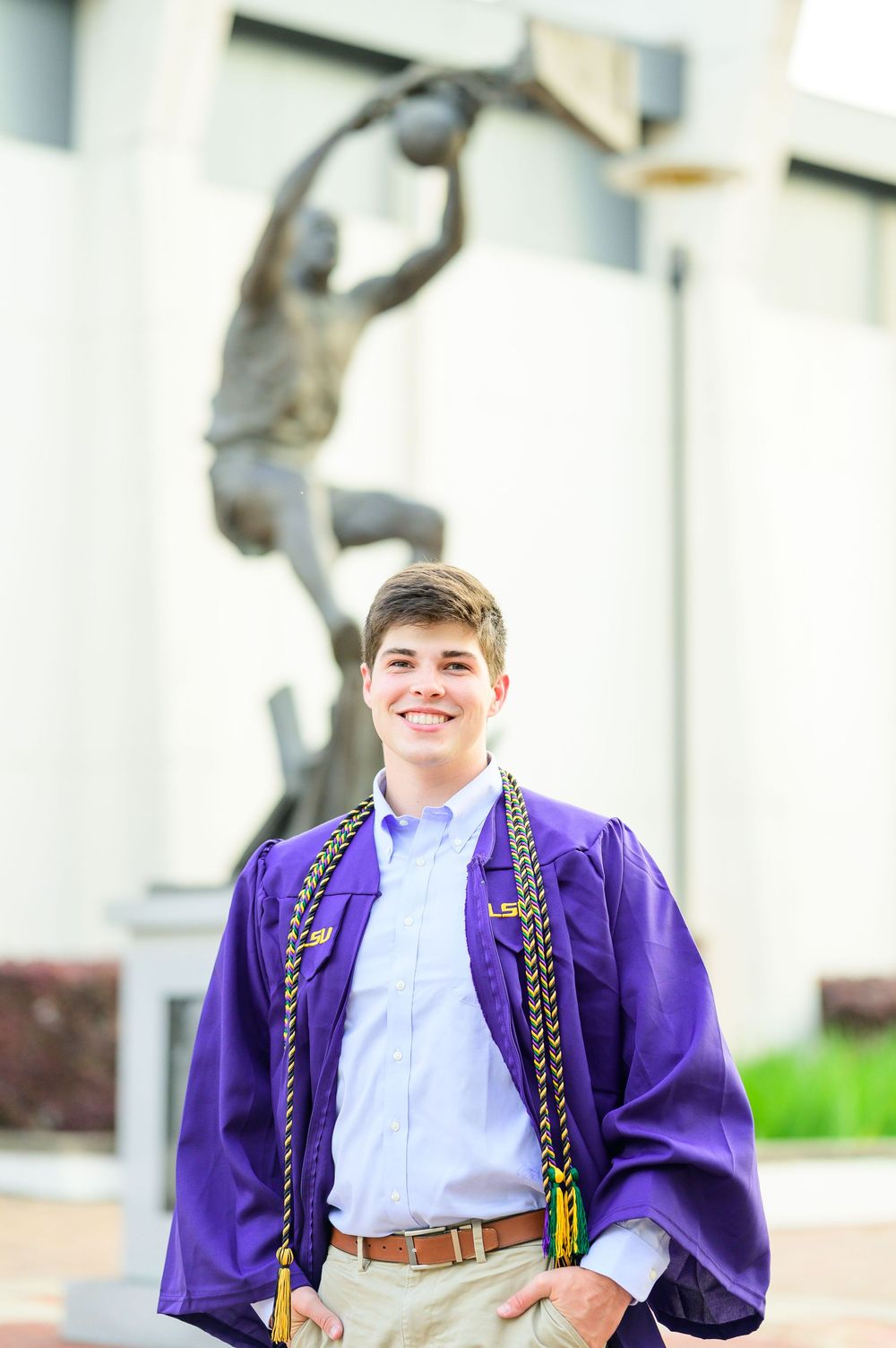 Graduate in purple LSU regalia standing in front of sports statue on campus.