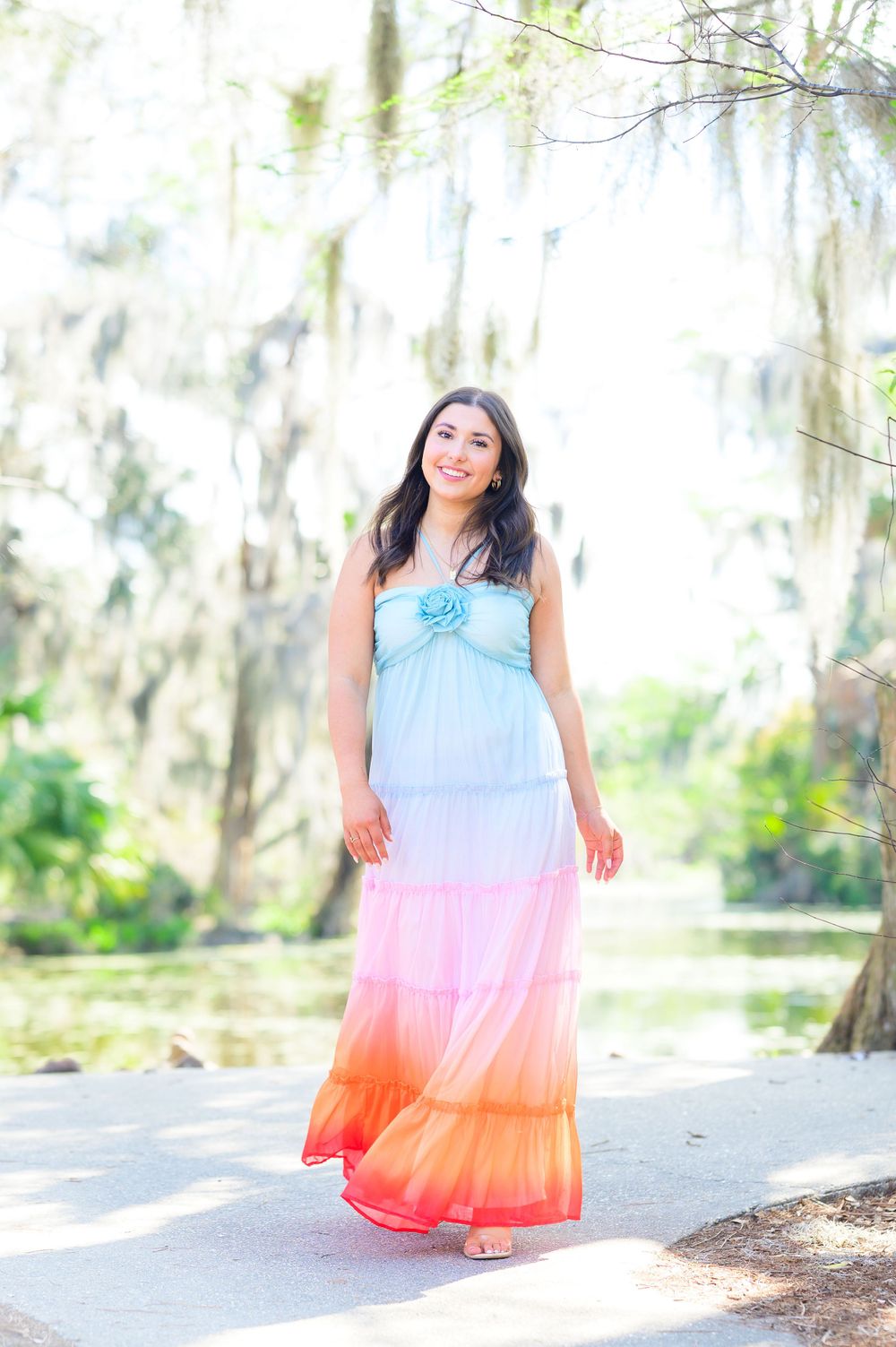 Model poses outdoors in a colorful ombre dress transitioning from light blue to coral pink against a natural backdrop.