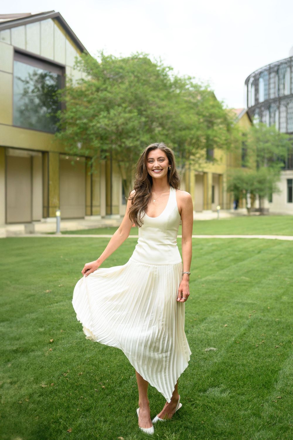 Young woman in white pleated midi dress smiling outdoors on green lawn with modern buildings in background.