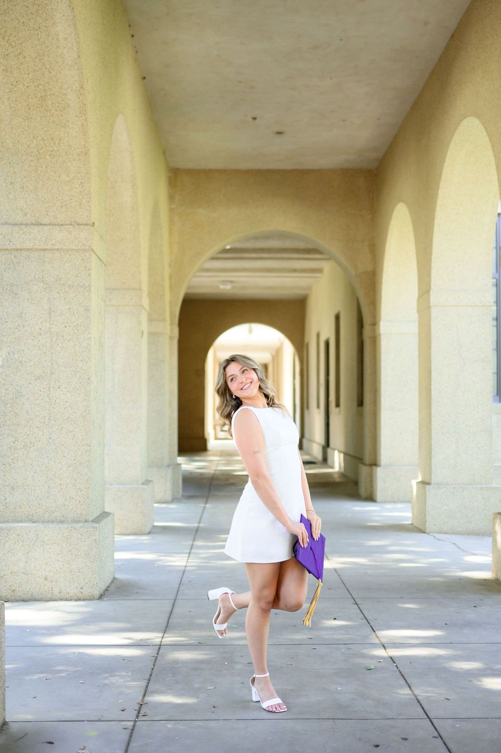 Young woman in white dress holding purple graduation cap poses under arched colonnade on sunny day.