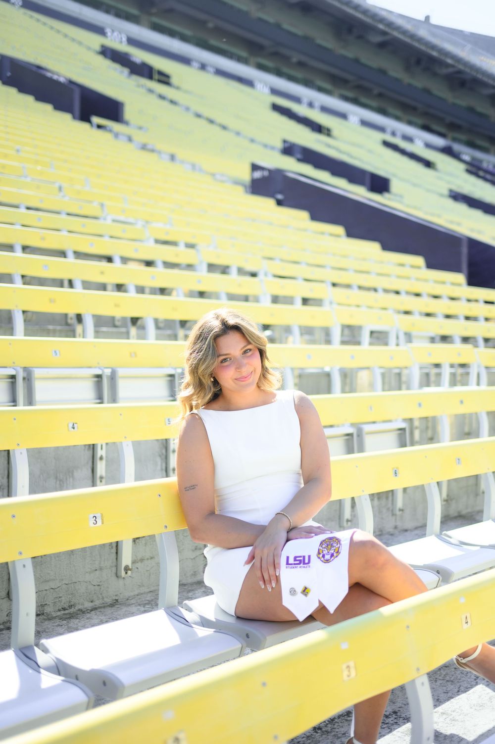 Young woman in white dress sitting in yellow stadium seats holding LSU graduation items.