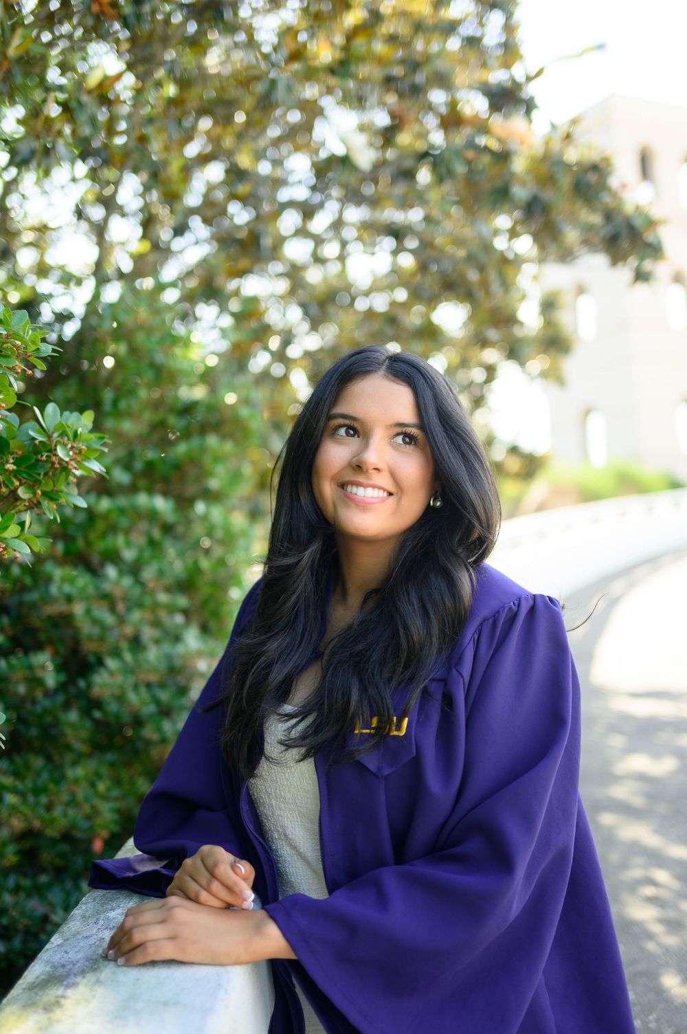 Smiling young woman in purple graduation gown leaning on stone railing outdoors with green trees in background.
