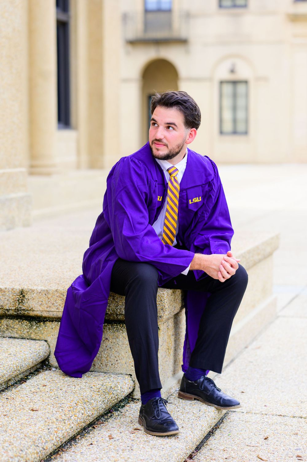 Graduate in LSU purple regalia sits thoughtfully on stone steps of campus building.