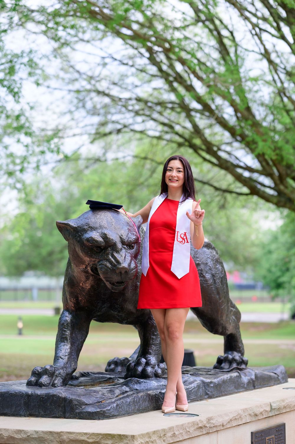 A smiling graduate in a red dress poses with a diploma on a bronze panther statue, wearing a USA sash outdoors.