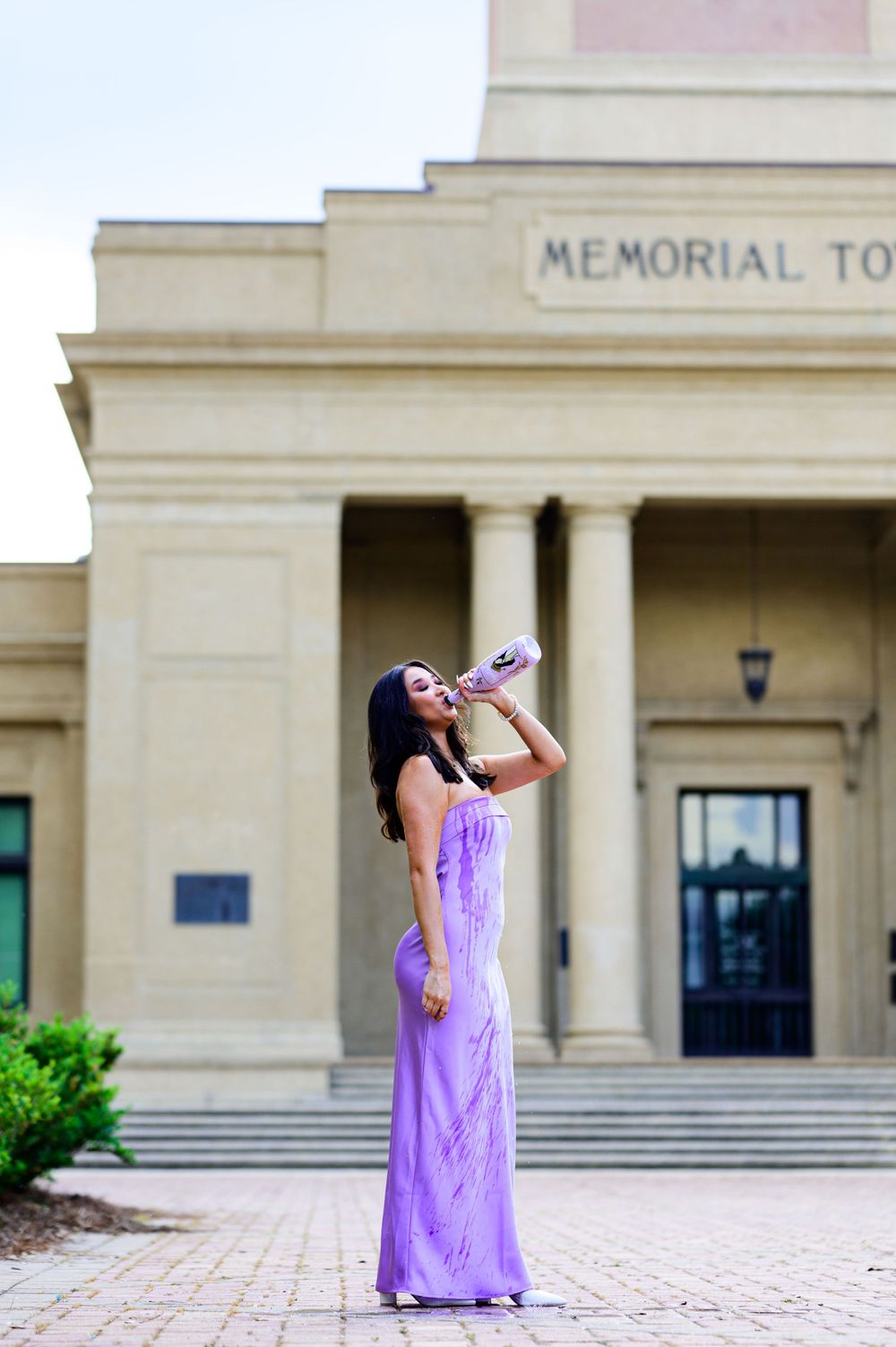 Person in purple formal dress poses with drink bottle outside Memorial Tower building.