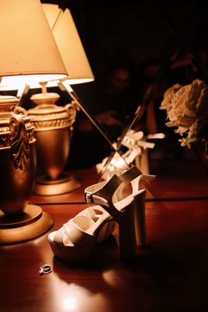 White bridal shoes displayed next to an elegant table lamp in warm lighting.