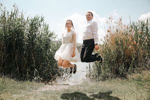 Couple jumps together in a grassy field wearing wedding attire.