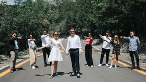 Wedding party walks in a line on a paved road surrounded by trees.