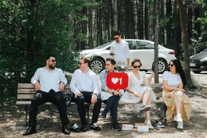Group relaxes on a bench in casual attire near parked cars in a forest setting.