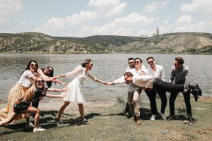 Wedding party poses playfully in a tug-of-war style photo by a scenic lake.