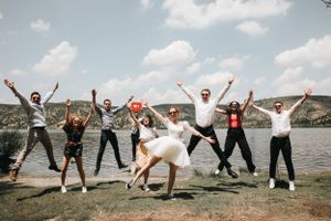 Wedding party jumps joyfully by a lake with mountains in the background on a sunny day.