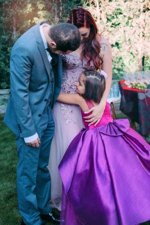 Family shares tender moment in garden setting with formal attire and purple dress.