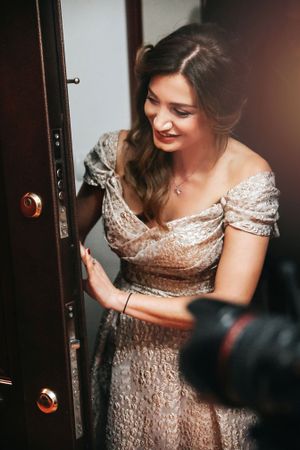 A woman in a sparkly champagne dress poses by a doorway with soft lighting.