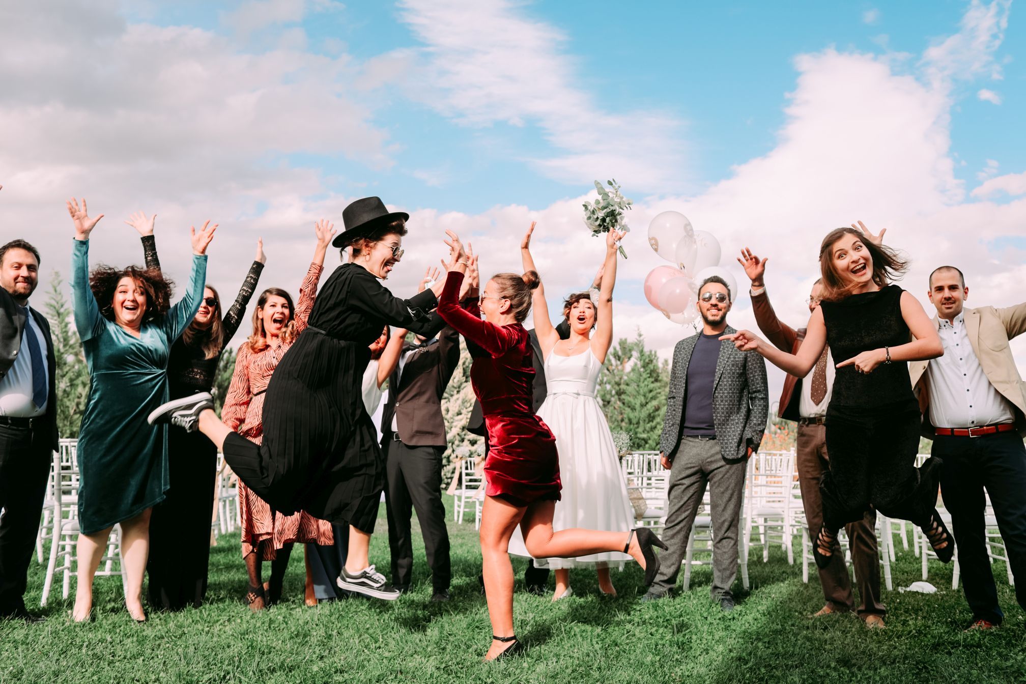 Wedding party jumps joyfully for group photo on sunny outdoor lawn.