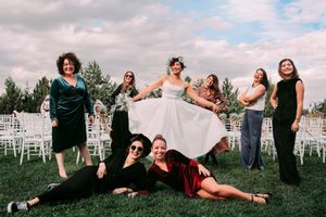 Wedding party poses playfully on grass with bridesmaids and white chairs in background.