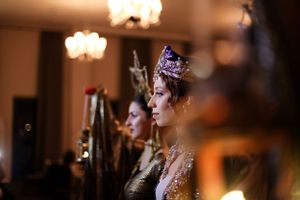 Side profile of performers with ornate crowns in atmospheric lighting at an evening event.