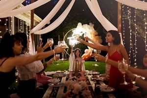 Outdoor celebration with string lights and white draping as guests toast across table.