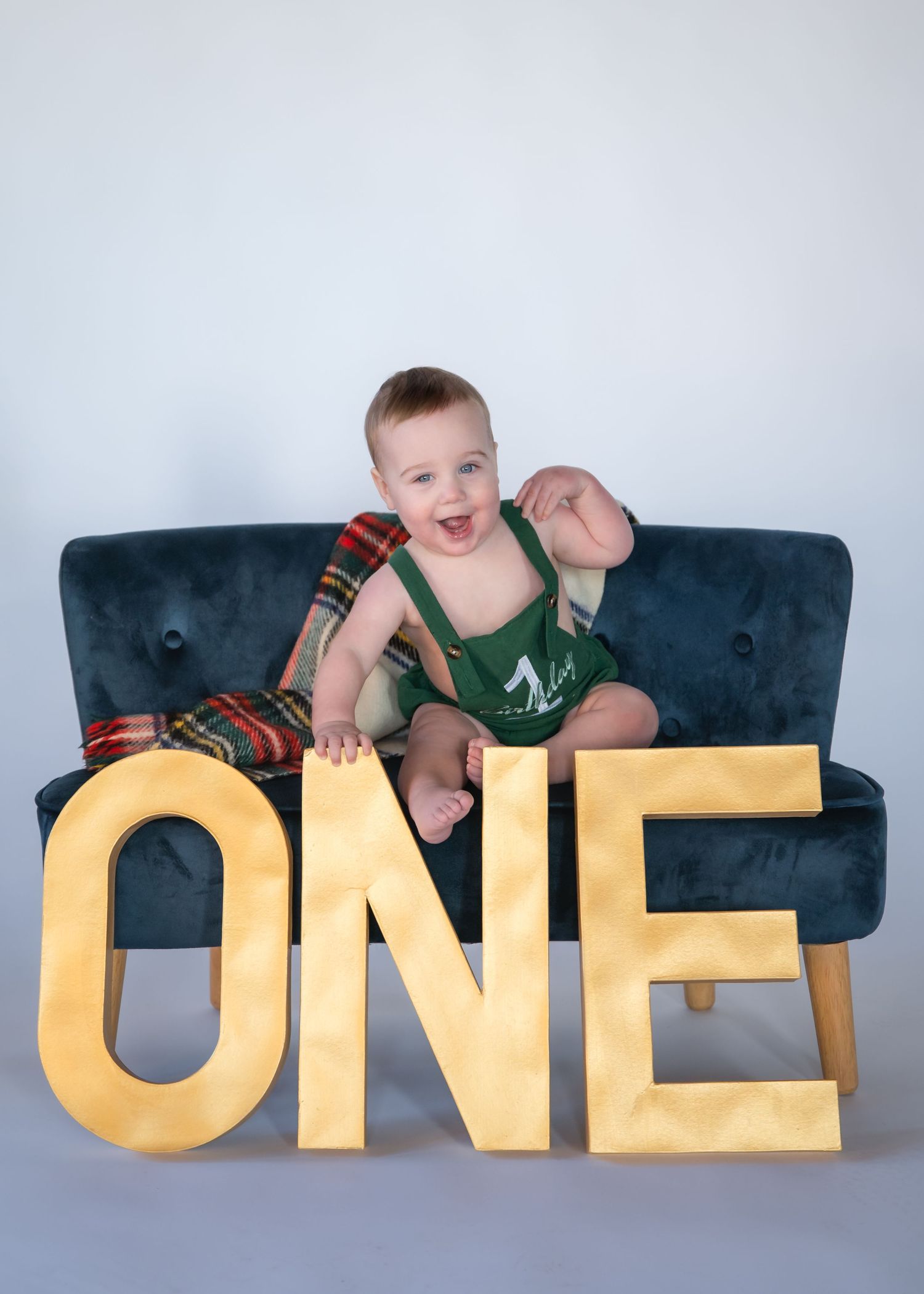 A smiling baby wearing green overalls and striped sleeves sits on a blue couch with large yellow letters spelling 'ONE'.