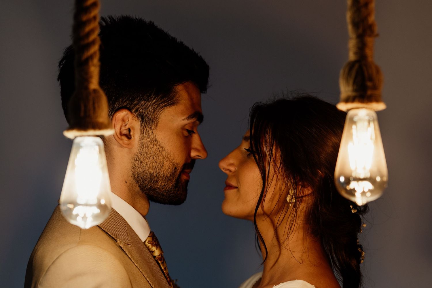 Romantic couple in wedding attire pose intimately between hanging vintage light bulbs.
