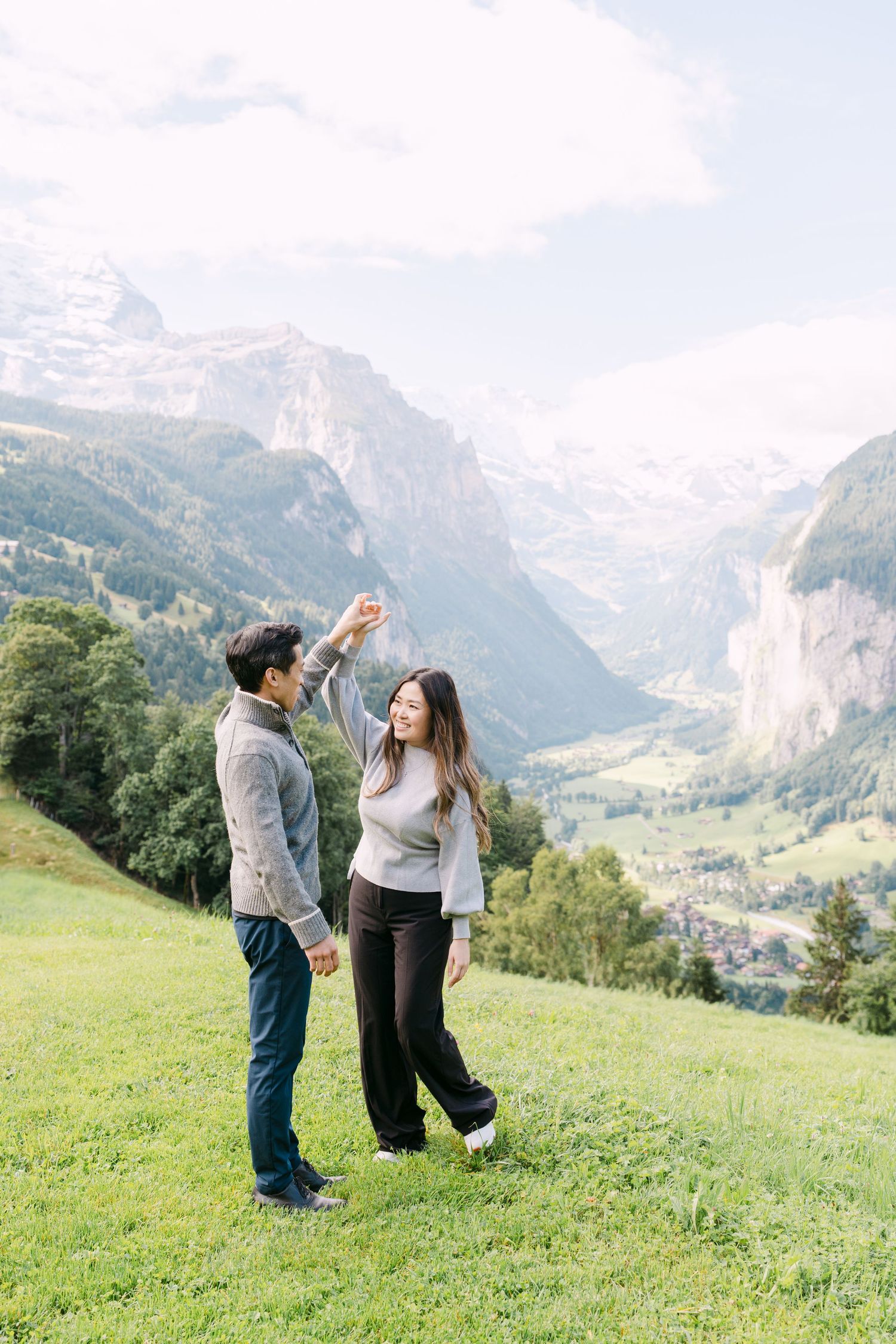 A couple sits together on a grassy hilltop overlooking a stunning mountain valley with snow-capped peaks in Lauterbrunnen, Switzerland.
