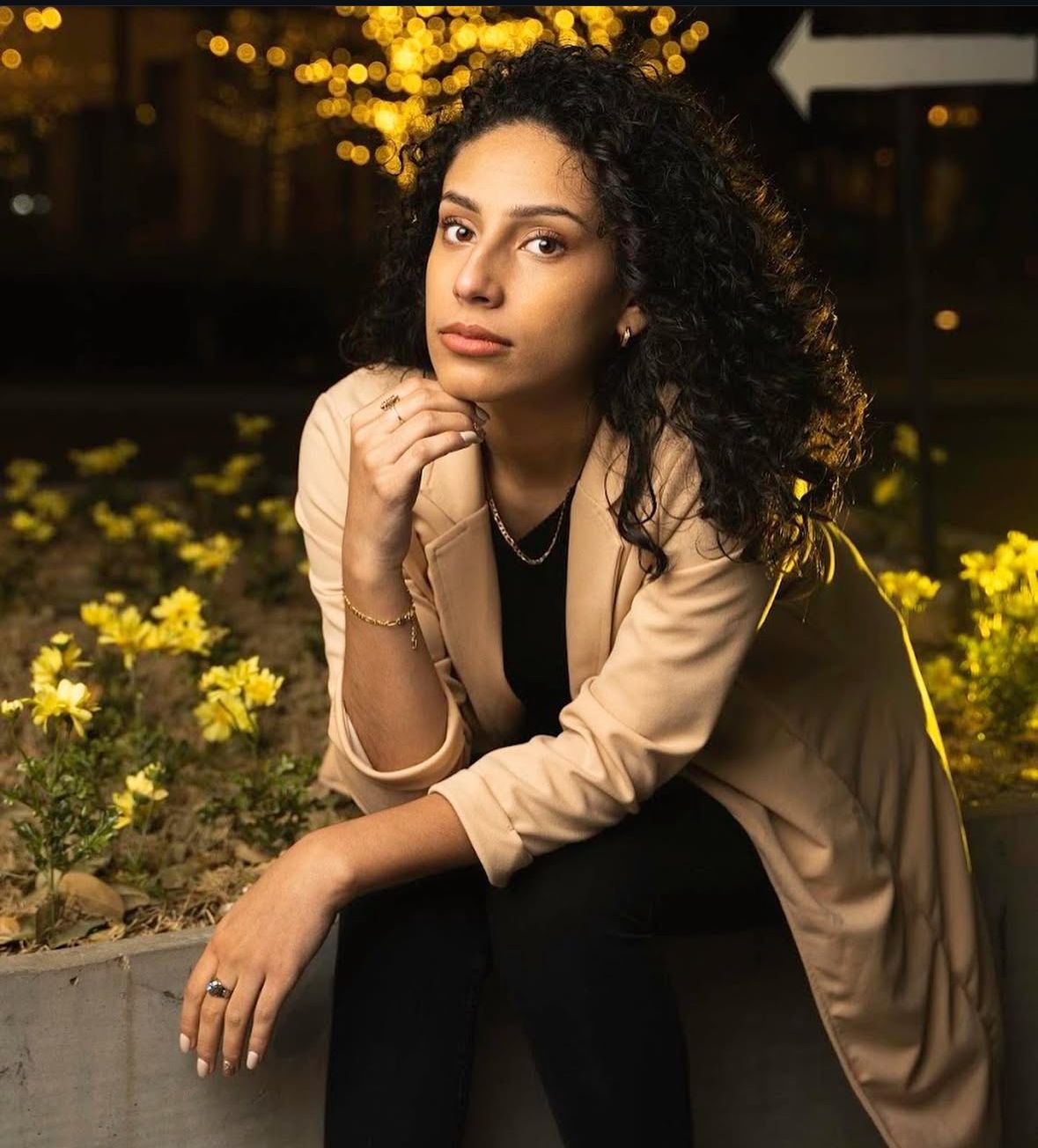 Portrait of someone in a beige blazer sitting thoughtfully by yellow flowers with bokeh lights in background at dusk.
