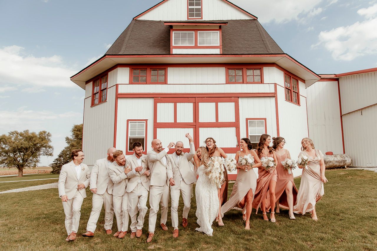 A wedding party in white suits and coral dresses poses together in front of a white and coral farmhouse on a sunny day.