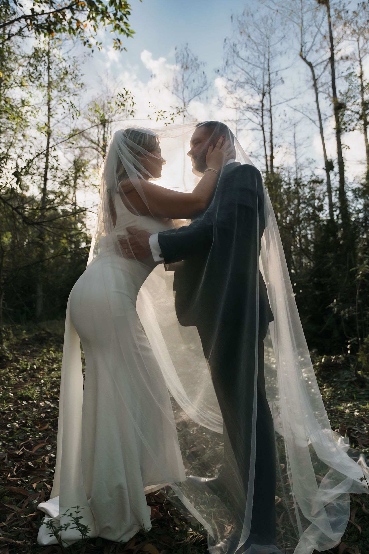 A romantic wedding photo silhouetted by sunlight through trees with flowing veils creating a dreamy outdoor atmosphere.
