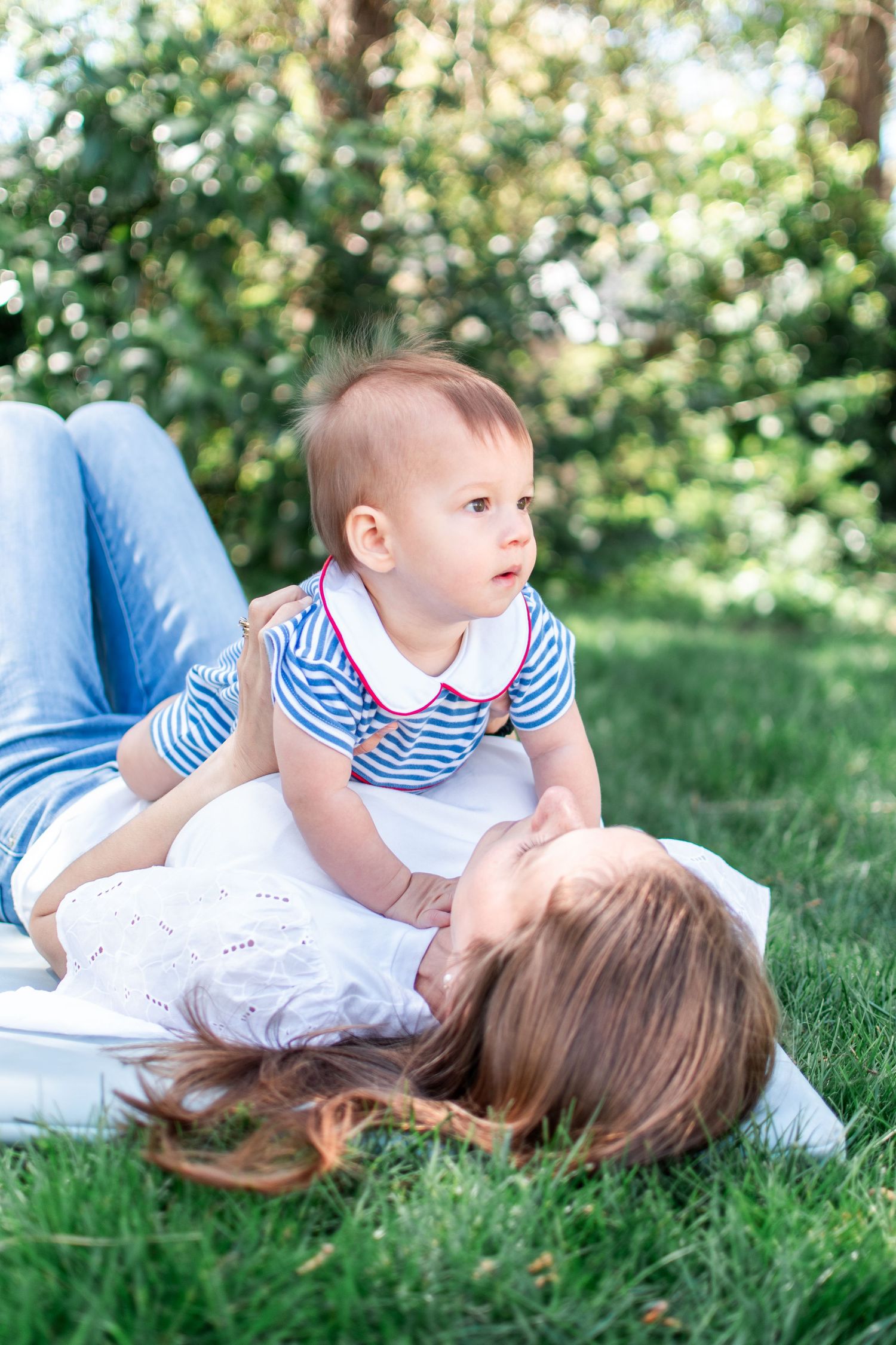 A baby in a striped shirt and white pants sits on their parent on lush green grass during a sunny day outdoors.