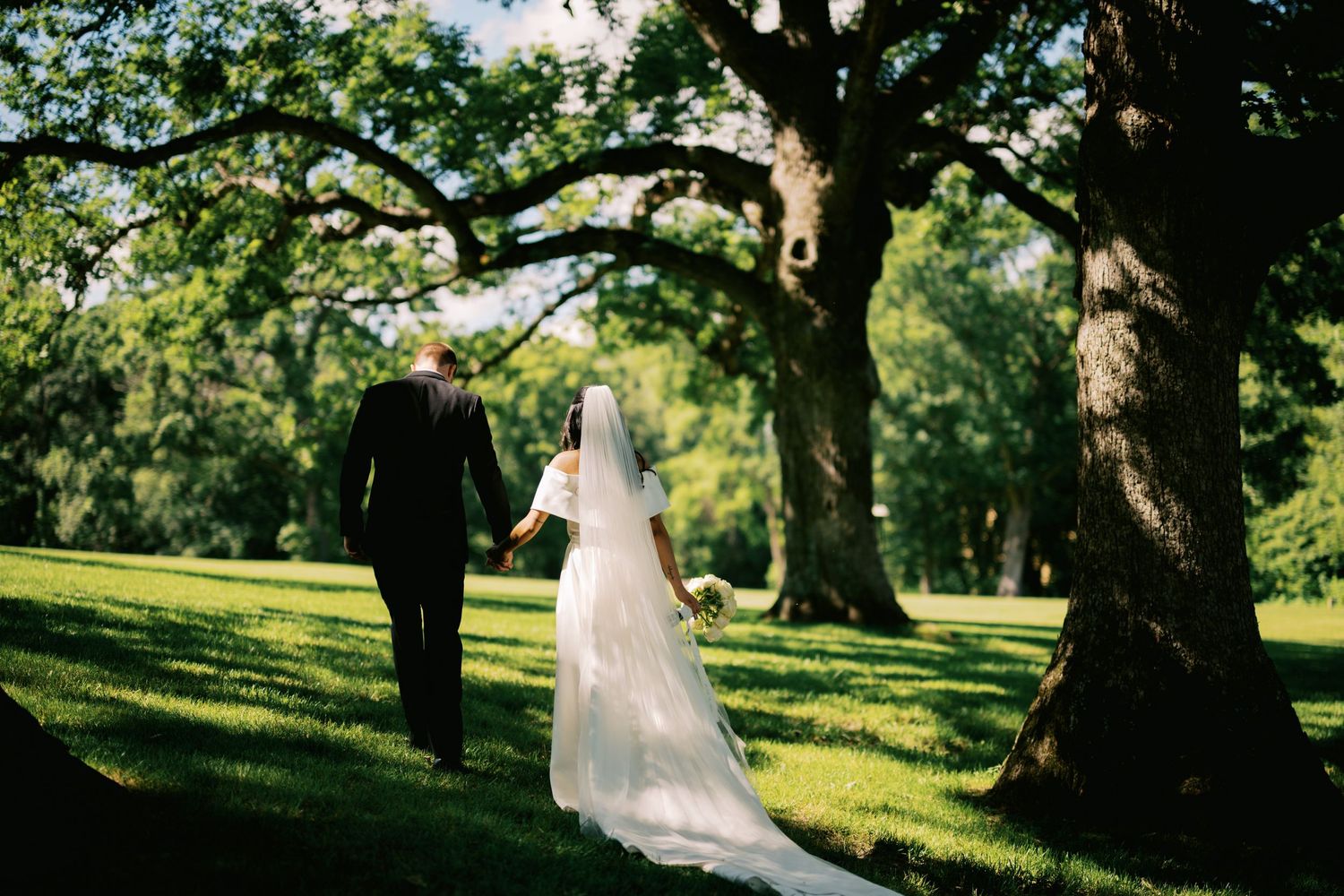 Two figures in wedding attire walk beneath towering oak trees on a sunny afternoon in a lush green park setting.